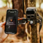 GSM Wildgame Innovations Encounter XT trail camera mounted to a tree beside a smartphone